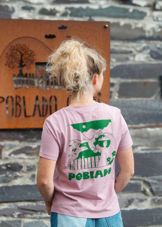 Person wearing a pink t-shirt with green graphics and text, standing in front of a stone wall and wooden sign.