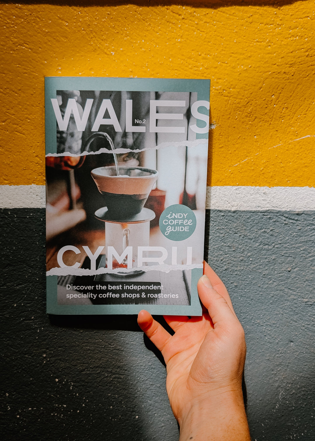 Hand holding a magazine titled 'Wales' against a yellow and gray wall.